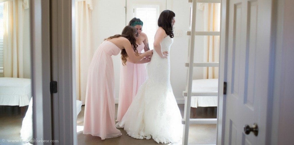 Bridesmaids tie the back of the bride's wedding gown while getting ready in the bridal suite.