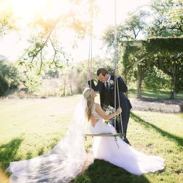 Bride sitting on swing looks at groom while her veil flows