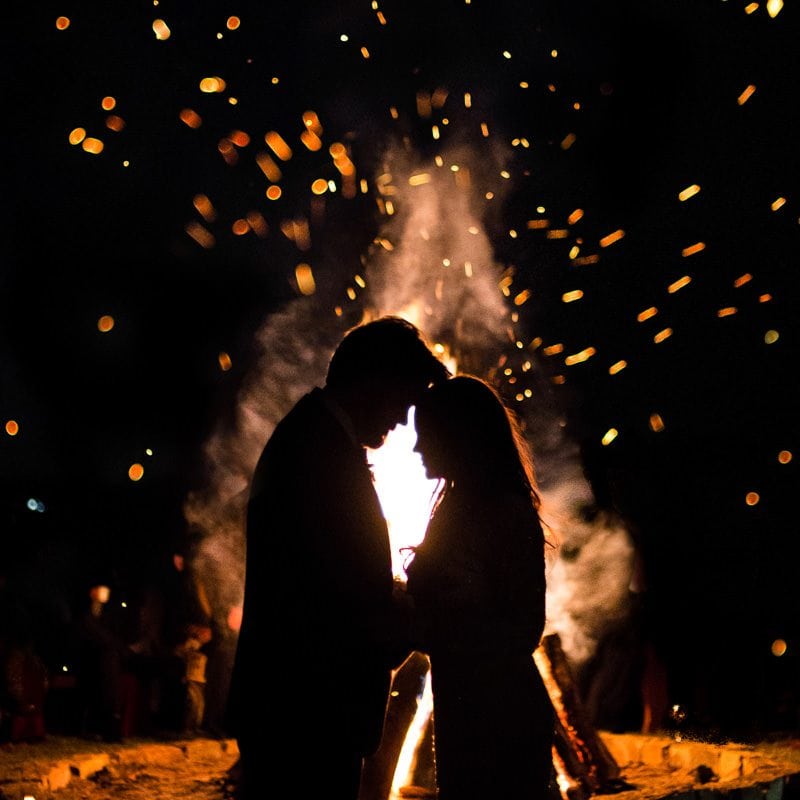 A silhouette of a couple in front of a bonfire tapping their foreheads together