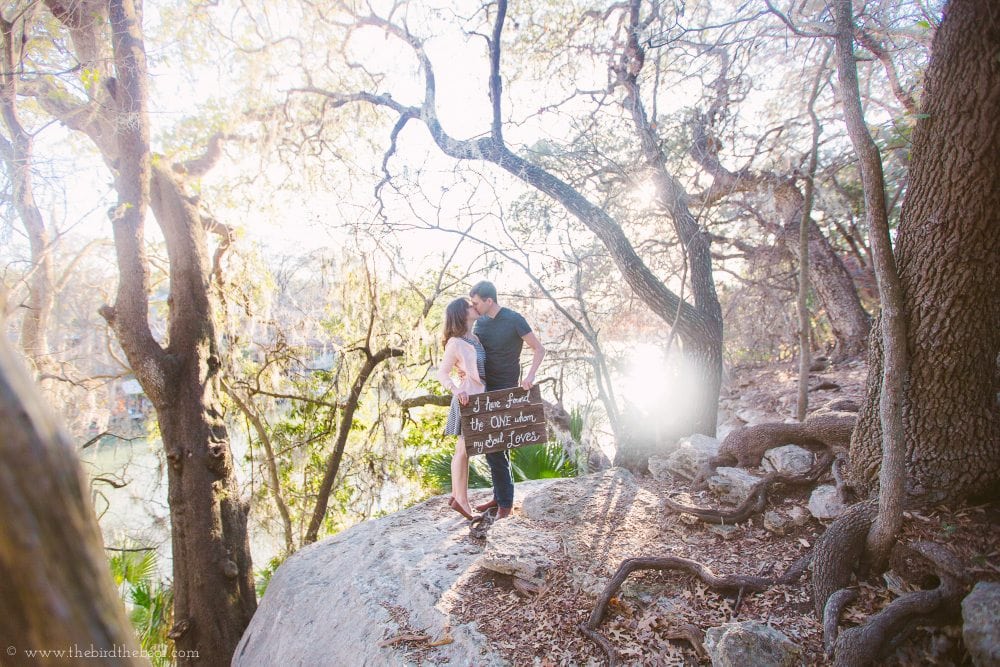 An engaged couple stands on the boulder overlooking Lake Austin in Mayfield Park.
