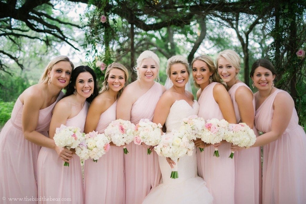 Bridesmaids wearing light pink, floor-length dresses with one shoulder strap.  
