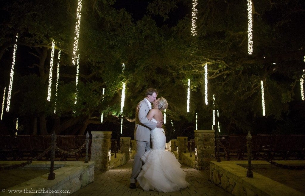 The bride and groom kiss under trees that are dripping with beautiful lights.