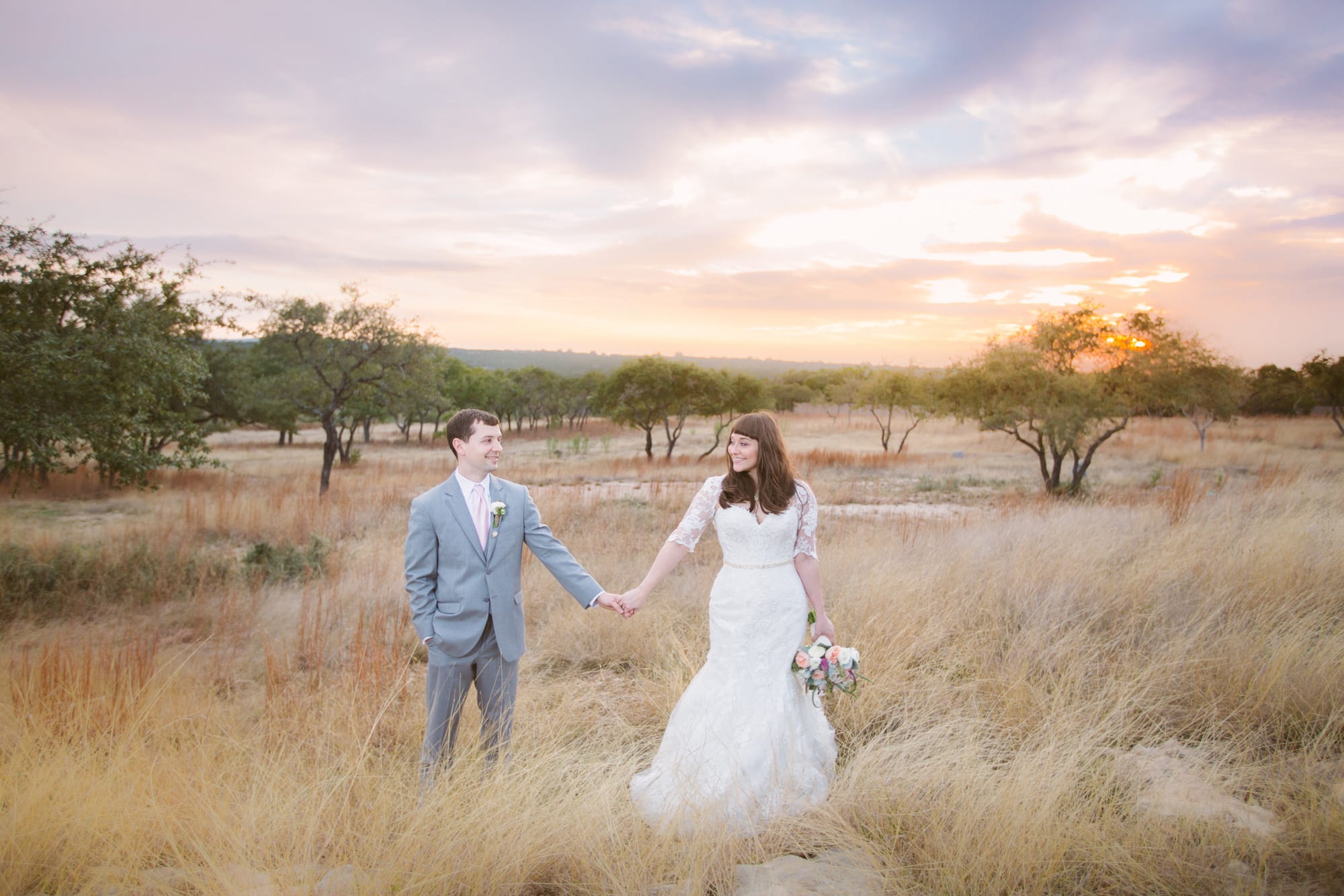 A bride and groom stand in field holding hands while photographers capture the romance